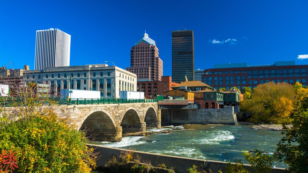 downtown Rochester skyline i bakgrunnen med en steinbro, Genesee-elven og planter i forgrunnsbildet tatt i løpet av høsten