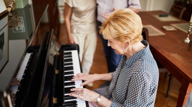 Senior kvinne som spiller piano for venner hjemme. Glade eldre kvinne som spiller piano med venner som lytter innendørs.