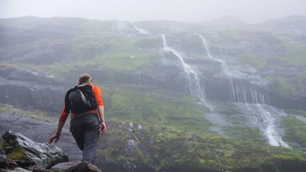 En kvinne med en liten ryggsekk og oransje stopper for en hvile mens vannet strømmer nedover fjellet i nærheten på Carrauntoohil i Killarney, Kerry i Irland