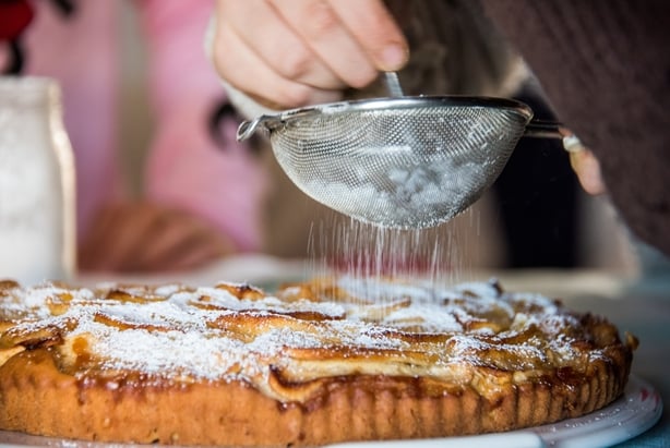 NÃ¦rbilde av et barn hjulpet av bestemoren Sifting Pulvered Sugar ved hjelp av en sil over en hjemmelaget eplebehandling