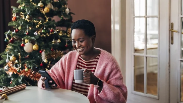 Afro-amerikansk kvinne nyter juleferien, drikker kaffe og surfer på sosiale medier på en mobiltelefon.