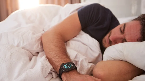 Mann sover i sengen med smartklokke mens solen bryter gjennom gardiner