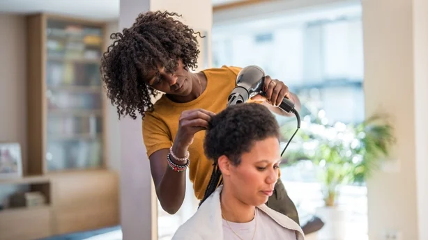 En middels voksen svart kvinne fletter håret til en kvinne av blandet rase i en koselig hjemmesetting.