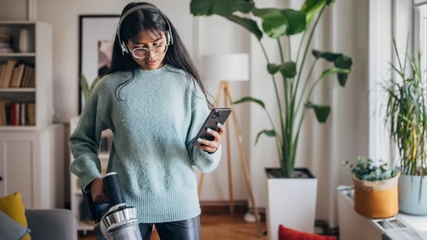En ung kvinne holder en trådløs støvsuger i den ene hånden og en smarttelefon i den andre i en koselig, plantefylt stue