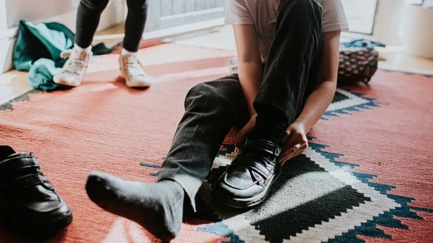 En ung gutt sitter på gulvet i et hjemlig miljø. Han drar på seg borrelås som fester svarte skolesko før han forlater huset for å starte skoledagen.