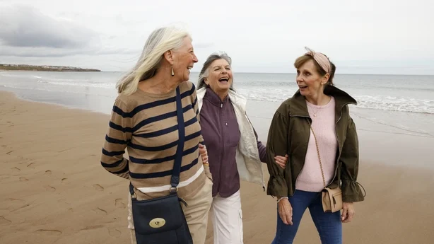 Tre eldre kvinnelige venner som går langs en strand og nyter tiden sammen, smiler og samhandler i et avslappet, gledelig øyeblikk. Bildet fanger vennskap, vennskap og velvære, og gjenspeiler gledene ved sosial tilknytning og aktiv, positiv aldring senere i livet.