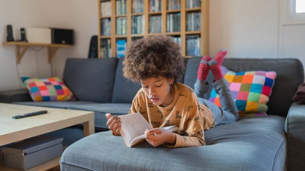 Afroamerikansk gutt leser en bok på sofaen. Forfra