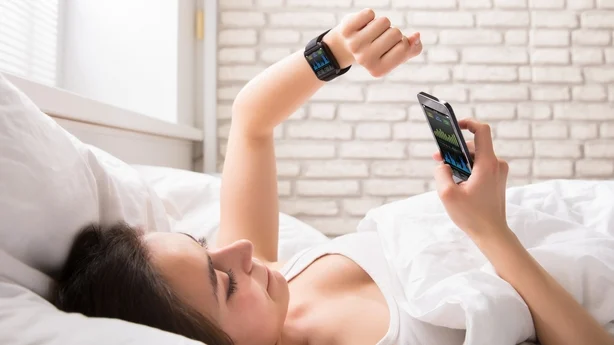 Nærbilde av en ung kvinne som ligger på sengen og synkroniserer smartklokke med mobiltelefon