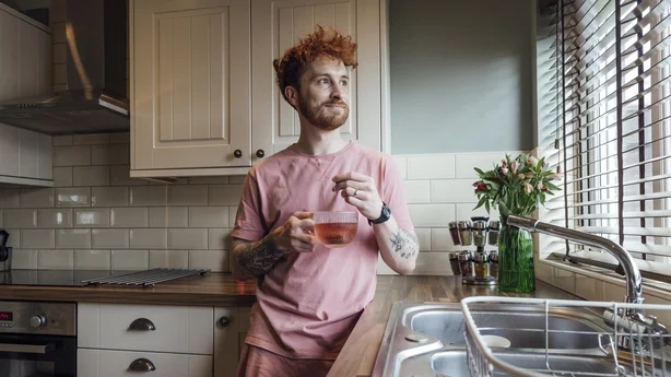 Tre fjerdedels lengde av en ung rødhåret mann som står på kjøkkenet sitt en morgen, han holder en kopp urtete og ser ut av vinduet i hjemmet sitt i County Durham, England. Han har på seg en uformell topp og shorts. Video er også tilgjengelig for dette scenariet.