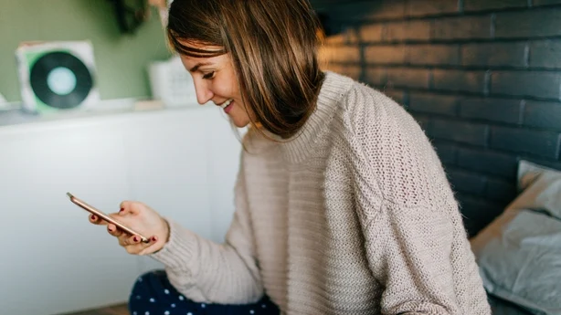 hvit kvinne sitter på en seng og ser på en telefon smilende