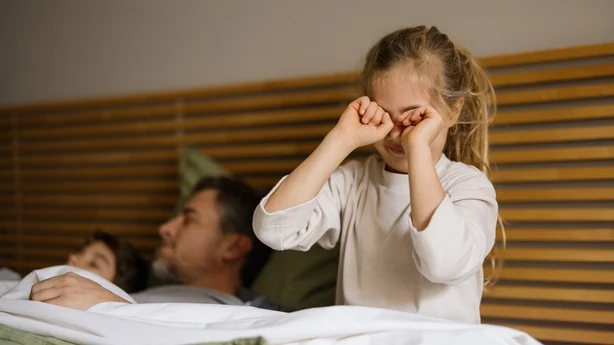 Jente som gnir seg i øynene mens hun våkner ved siden av sin sovende far og søsken i sengen, og starter dagen