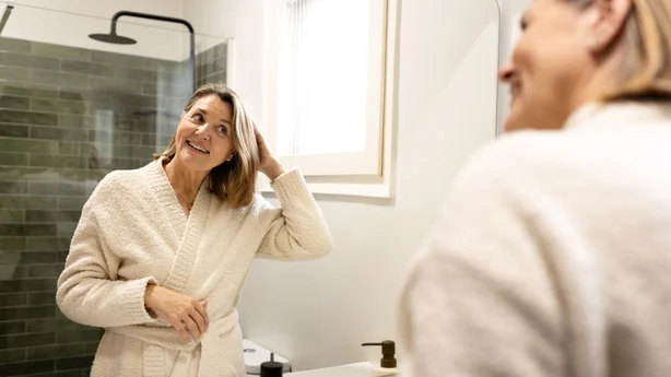 Smilende senior kvinne iført badekåpe, ser på seg selv i speilet og fikser håret på badet
